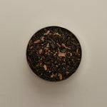 Overhead view of loose herbal tea blend in a round tin, featuring dried tea leaves on a beige background.
