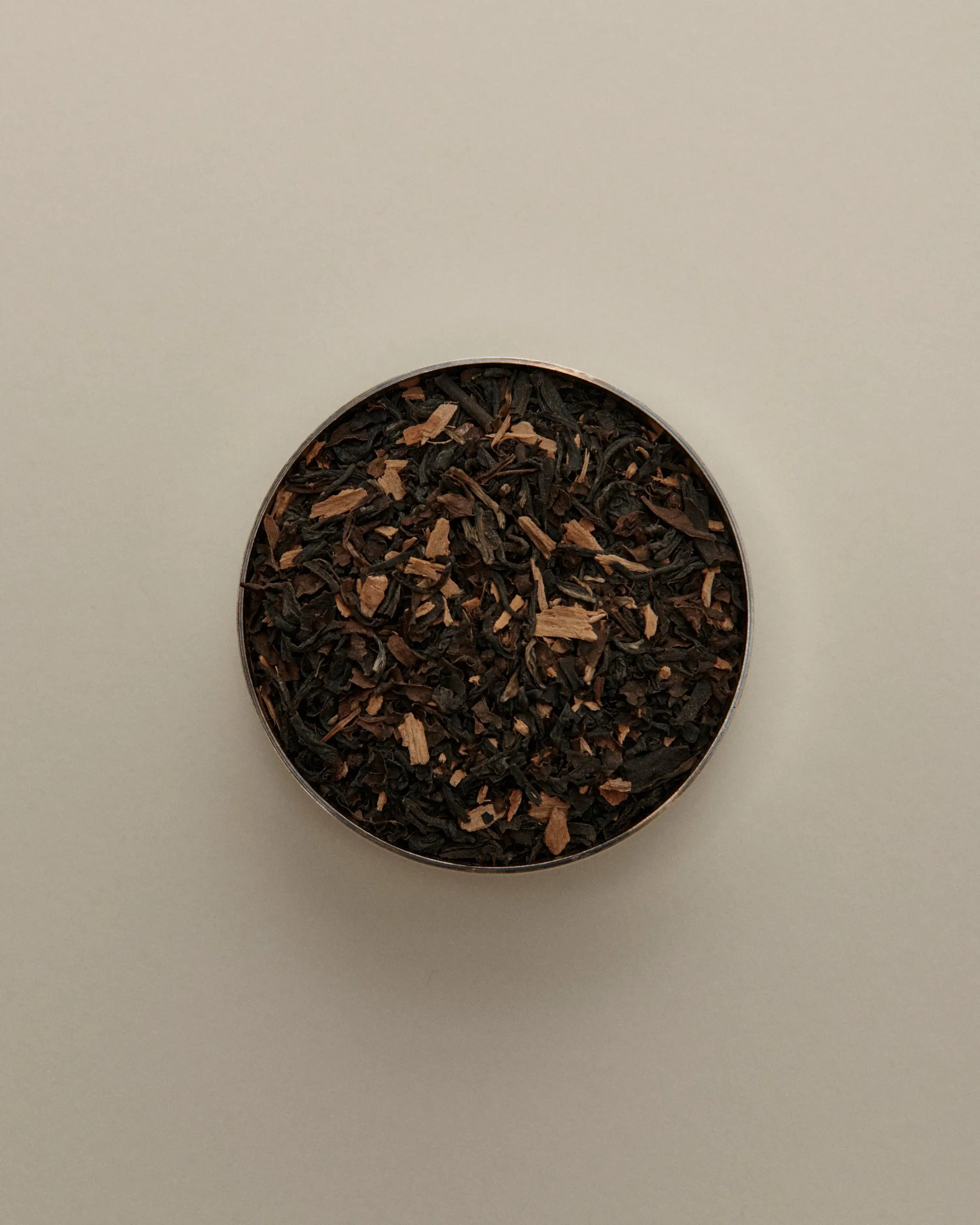 Overhead view of loose herbal tea blend in a round tin, featuring dried tea leaves on a beige background.