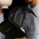 A person holds a white Forgrave "005 Mint Mantra Herbal Blend" box, reading the back label while holding a teacup, almost ready to drink.