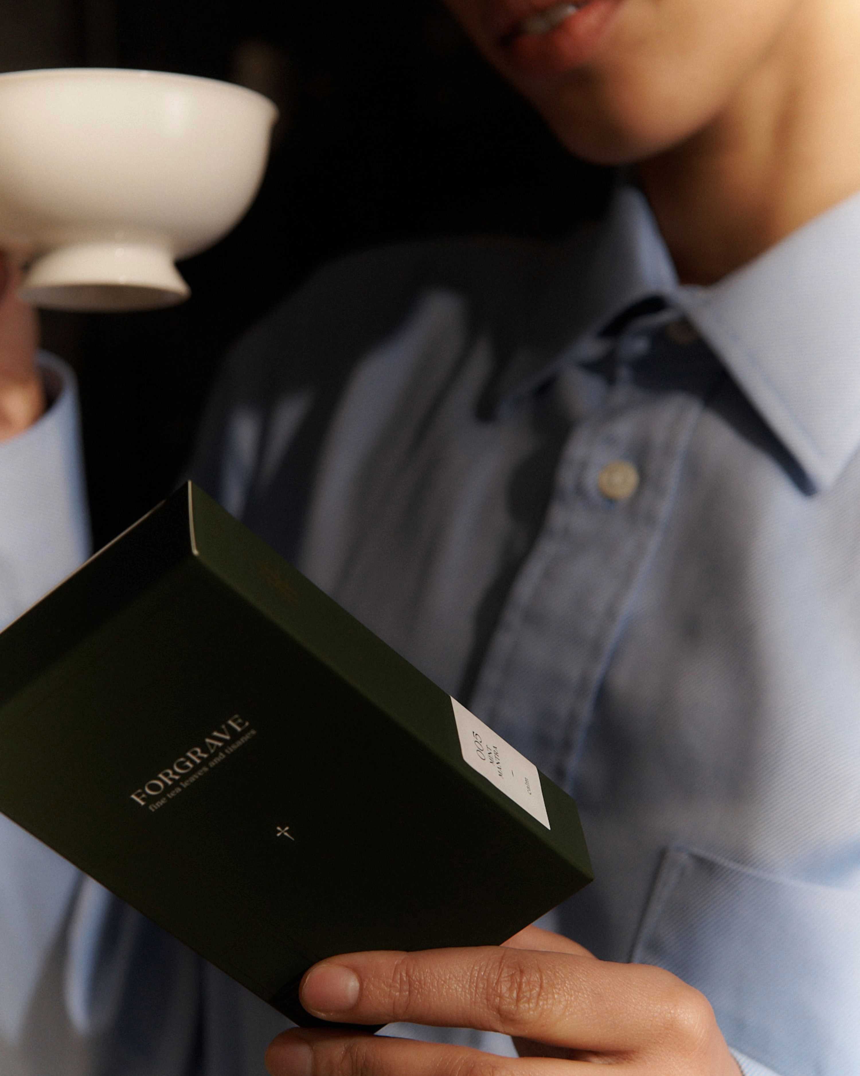A person holds a white Forgrave "005 Mint Mantra Herbal Blend" box, reading the back label while holding a teacup, almost ready to drink.