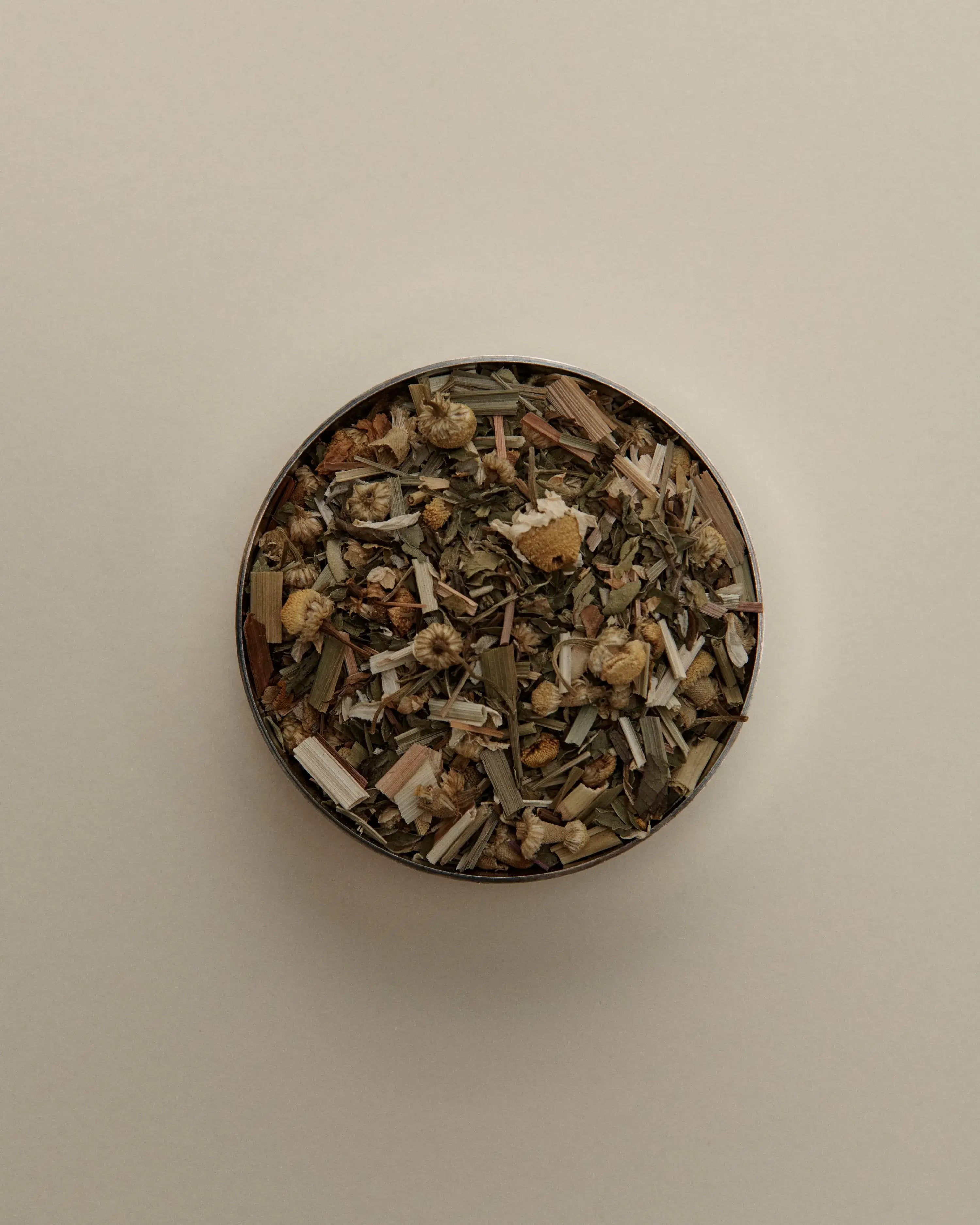 Overhead view of loose herbal tea blend in a round tin, featuring dried tea leaves on a beige background.