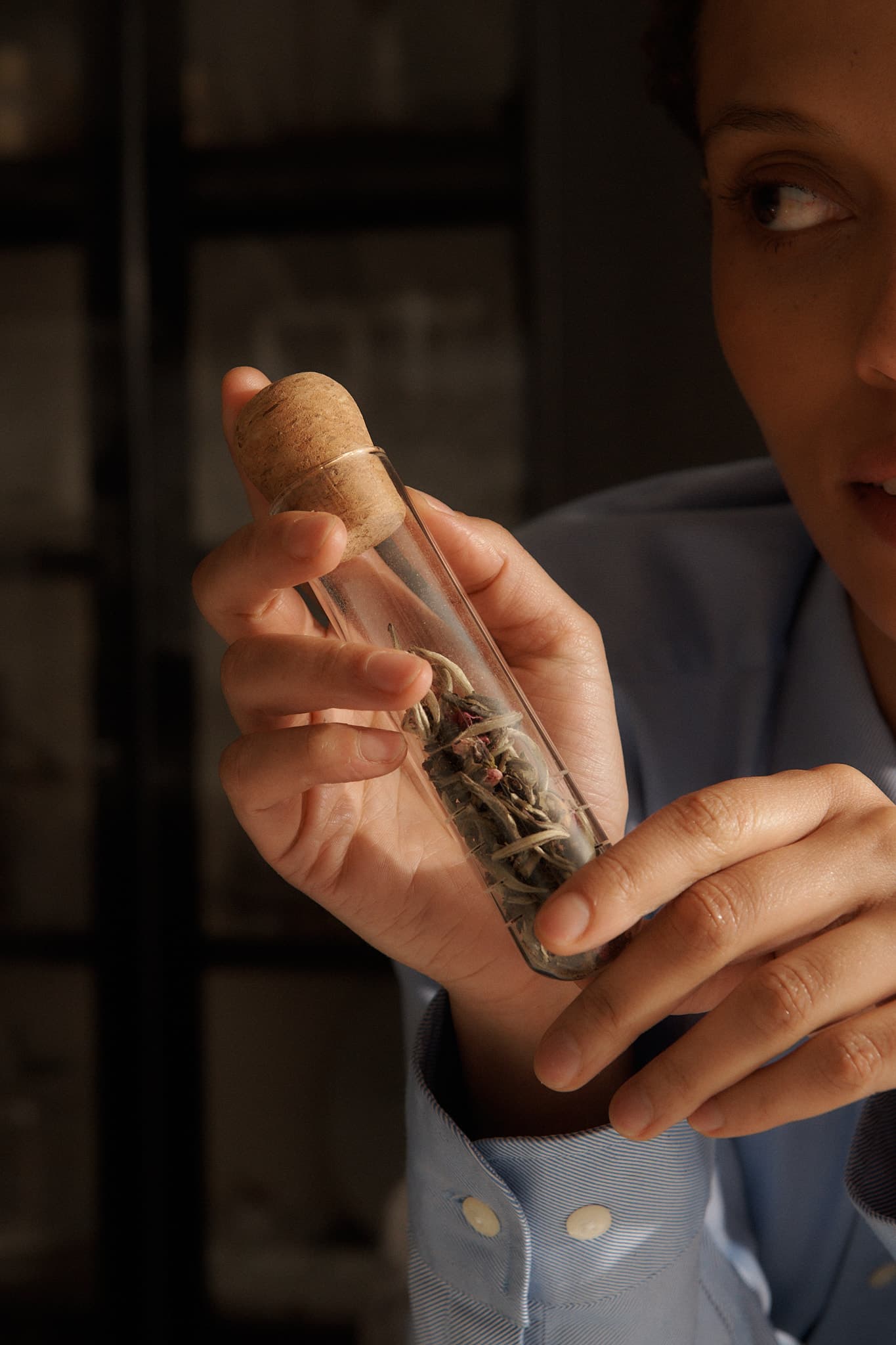 Hand holding a glass tea infuser tube with a cork lid, filled with loose tea leaves, against a dimly lit background.