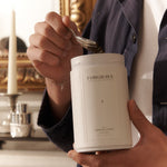 Person scooping tea from a white ‘FORGRAVE Smoking Jacket Green Blend’ canister, with a gold-framed mirror and candle in the background.