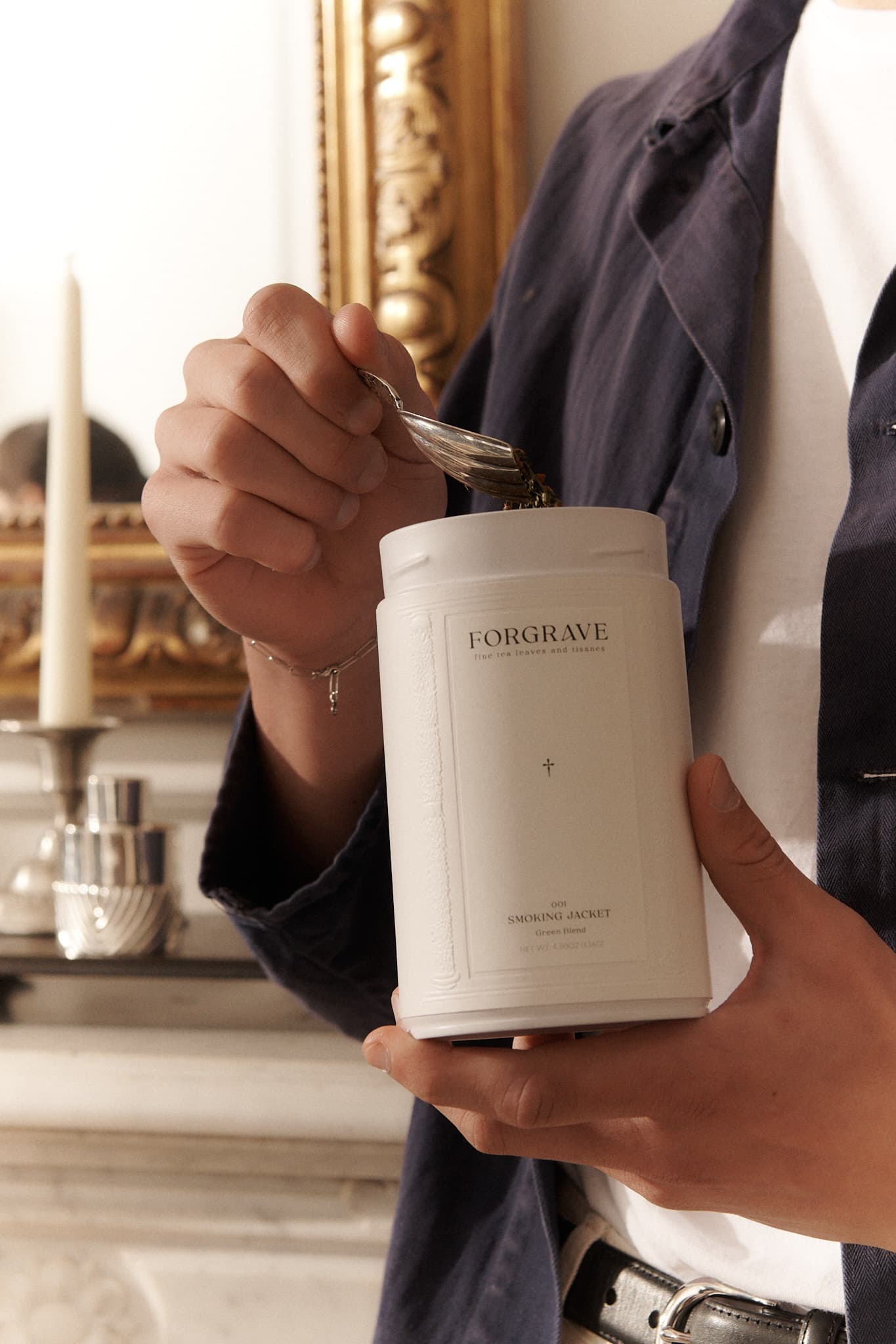 Person scooping tea from a white ‘FORGRAVE Smoking Jacket Green Blend’ canister, with a gold-framed mirror and candle in the background.