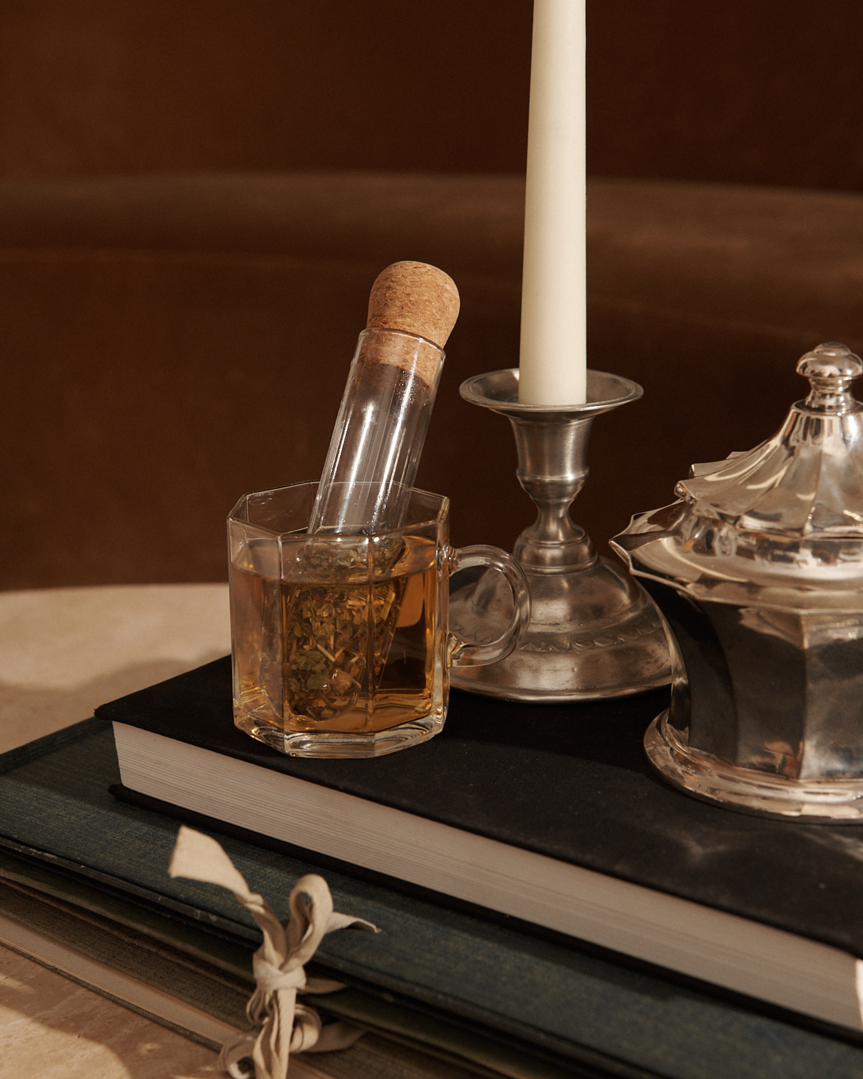 A glass mug of herbal tea with a corked glass infuser sits on stacked books beside a silver candlestick and sugar container.