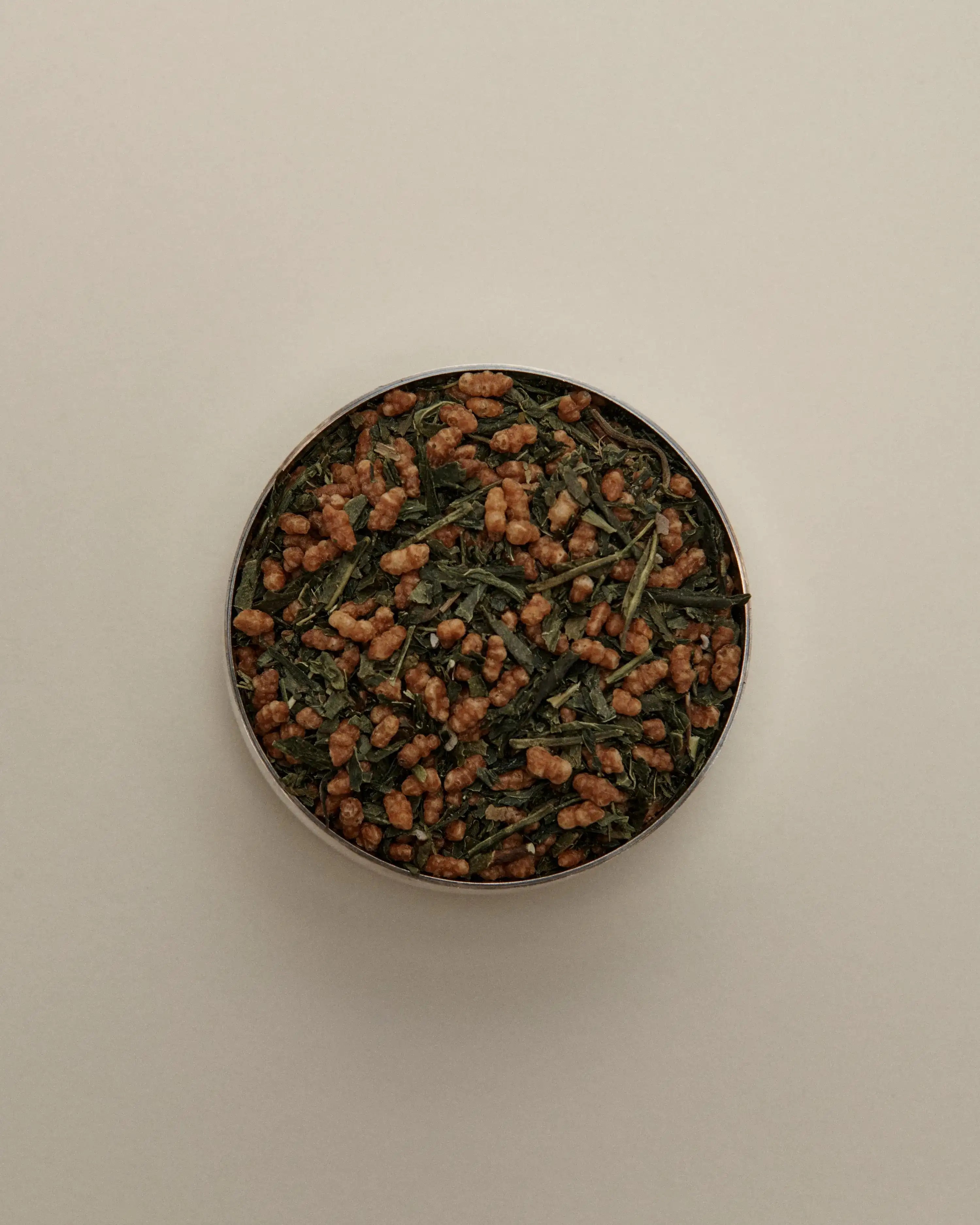 Overhead view of loose herbal tea blend in a round tin, featuring dried tea leaves on a beige background.
