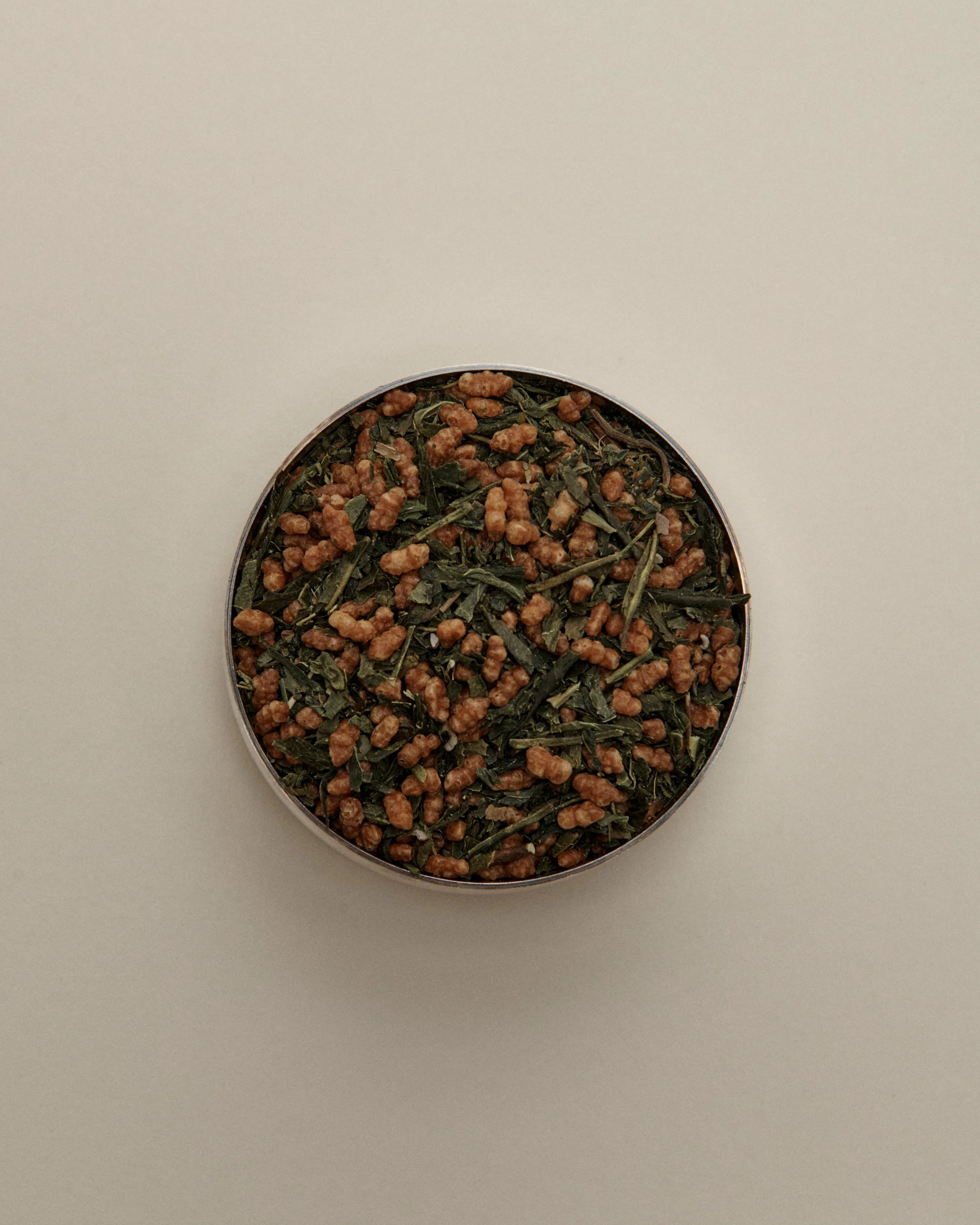 Overhead view of loose herbal tea blend in a round tin, featuring dried tea leaves on a beige background.
