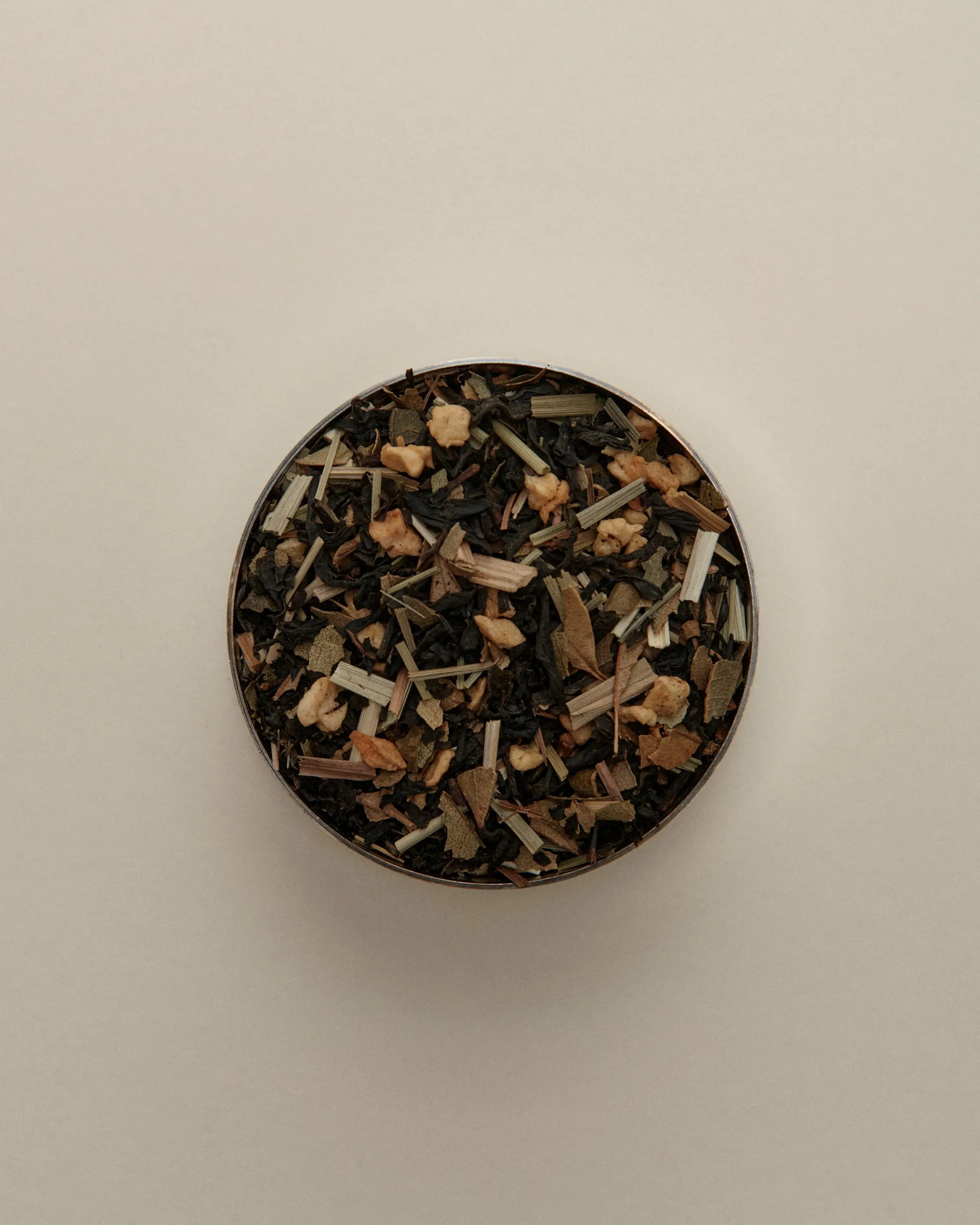 Overhead view of loose herbal tea blend in a round tin, featuring dried tea leaves beige background.