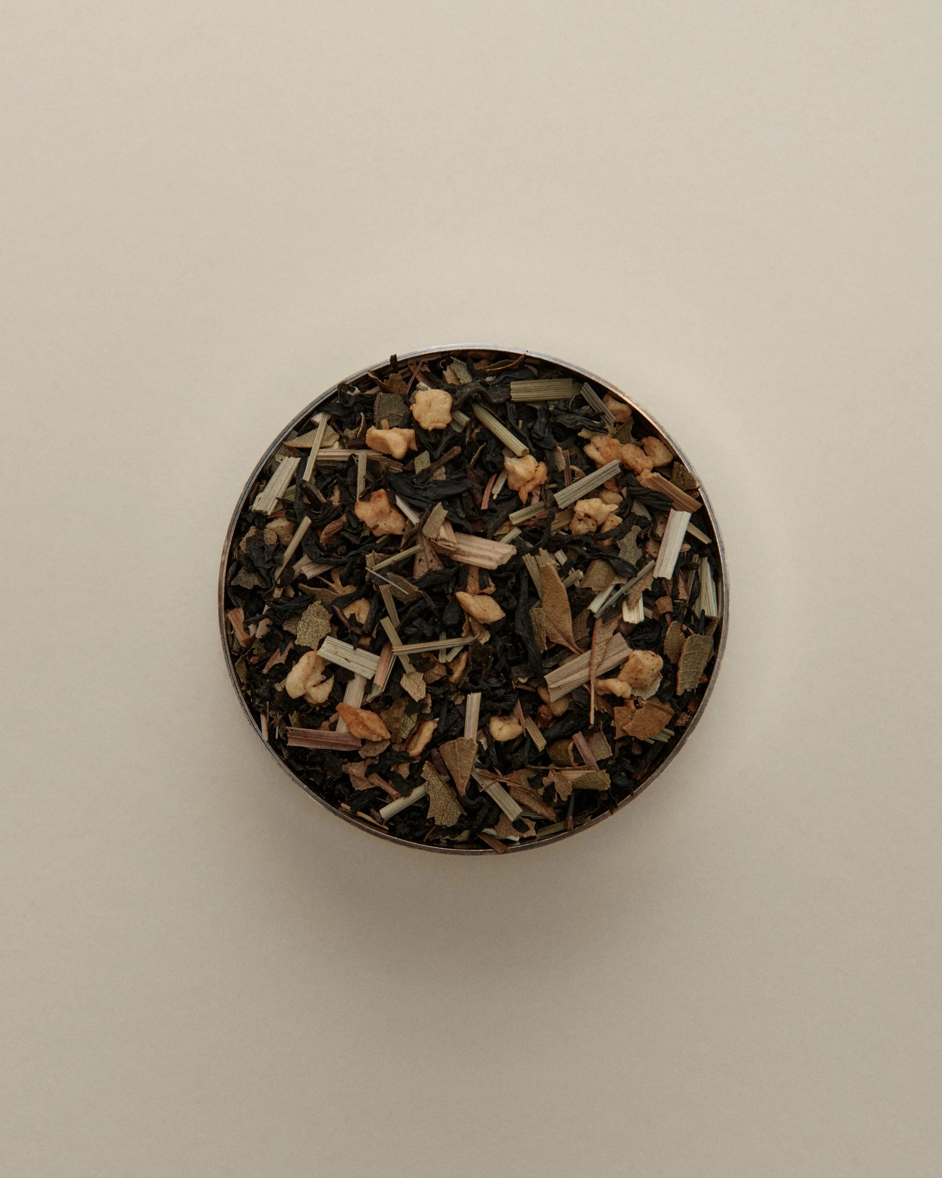 Overhead view of loose herbal tea blend in a round tin, featuring dried tea leaves beige background.