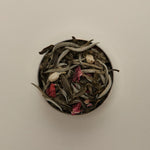 Overhead view of loose herbal tea blend in a round tin, featuring dried tea leaves on a beige background.