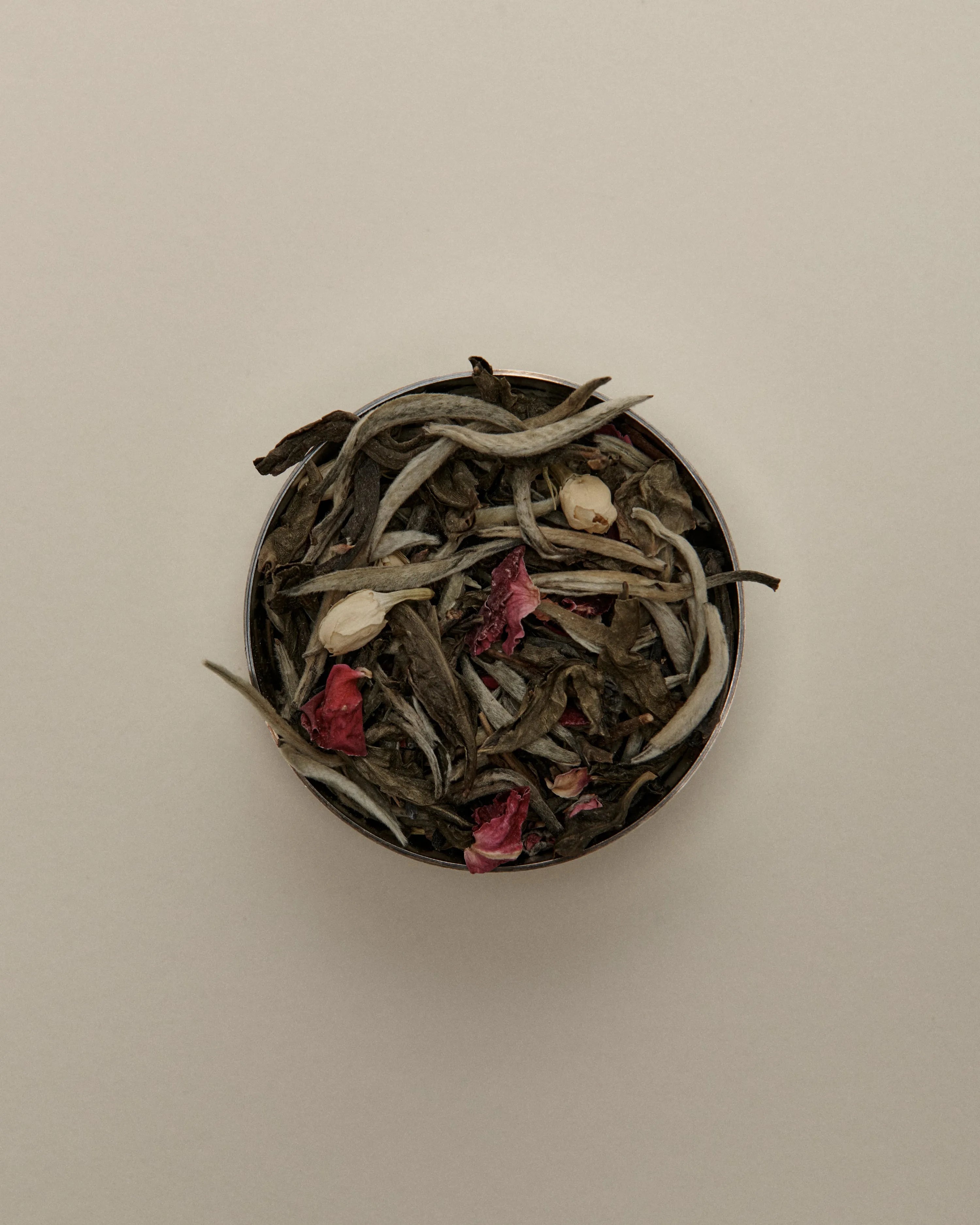 Overhead view of loose herbal tea blend in a round tin, featuring dried tea leaves on a beige background.