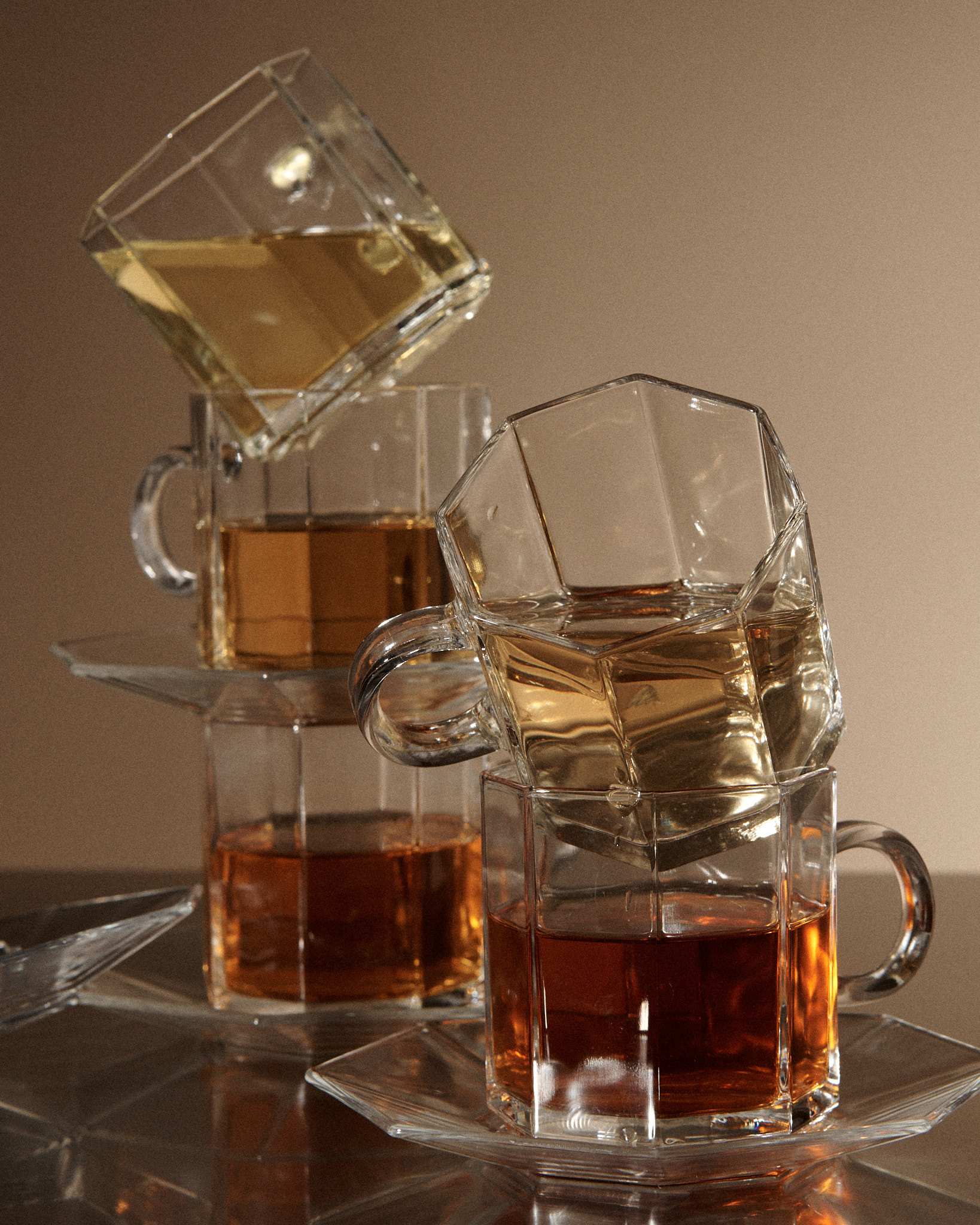 Stacked clear glass mugs filled with various shades of tea, reflecting warm light on a beige background.