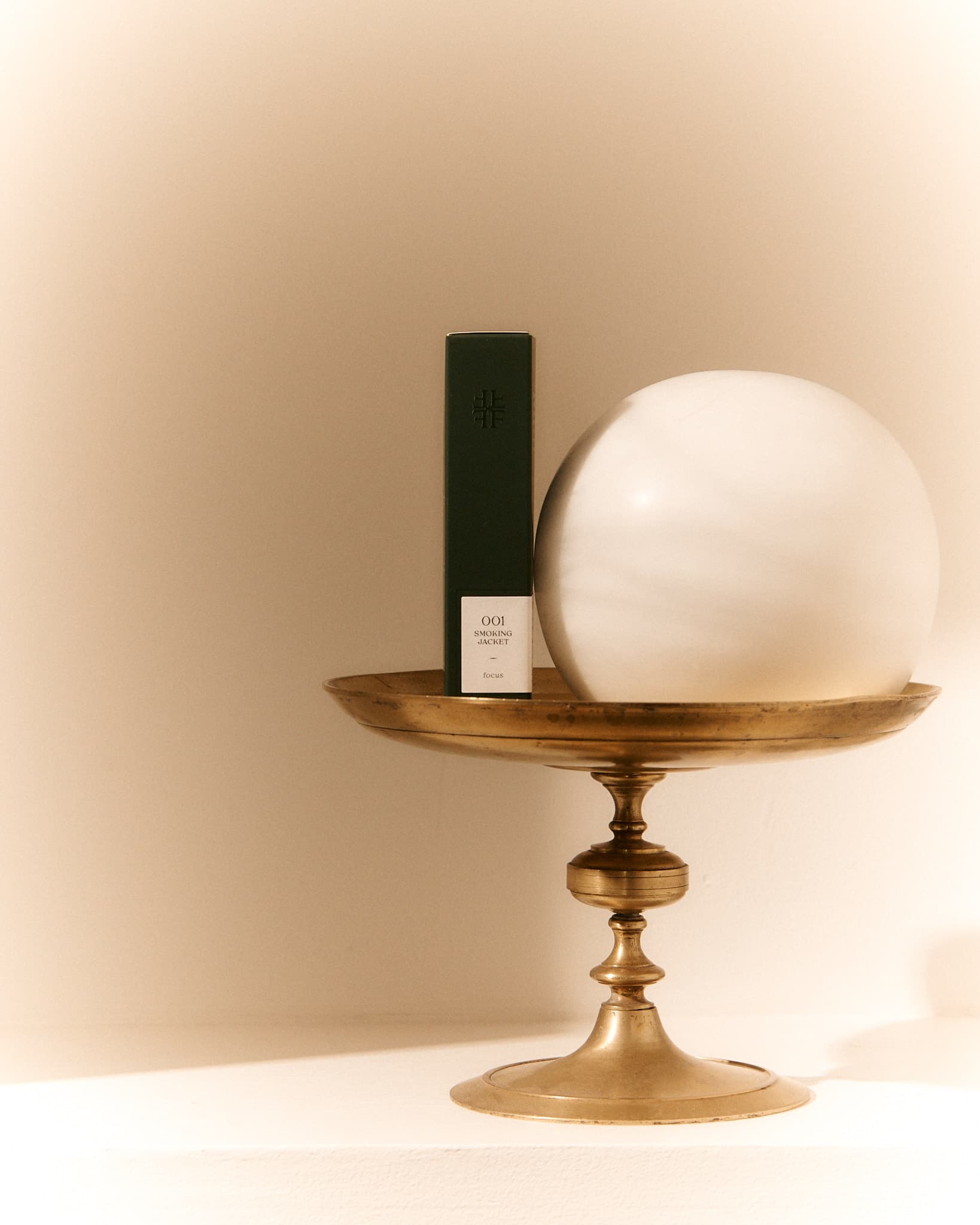 Dark green FORGRAVE ‘001 Smoking Jacket – Focus’ tea box displayed on a brass pedestal with a white marble orb beside it.