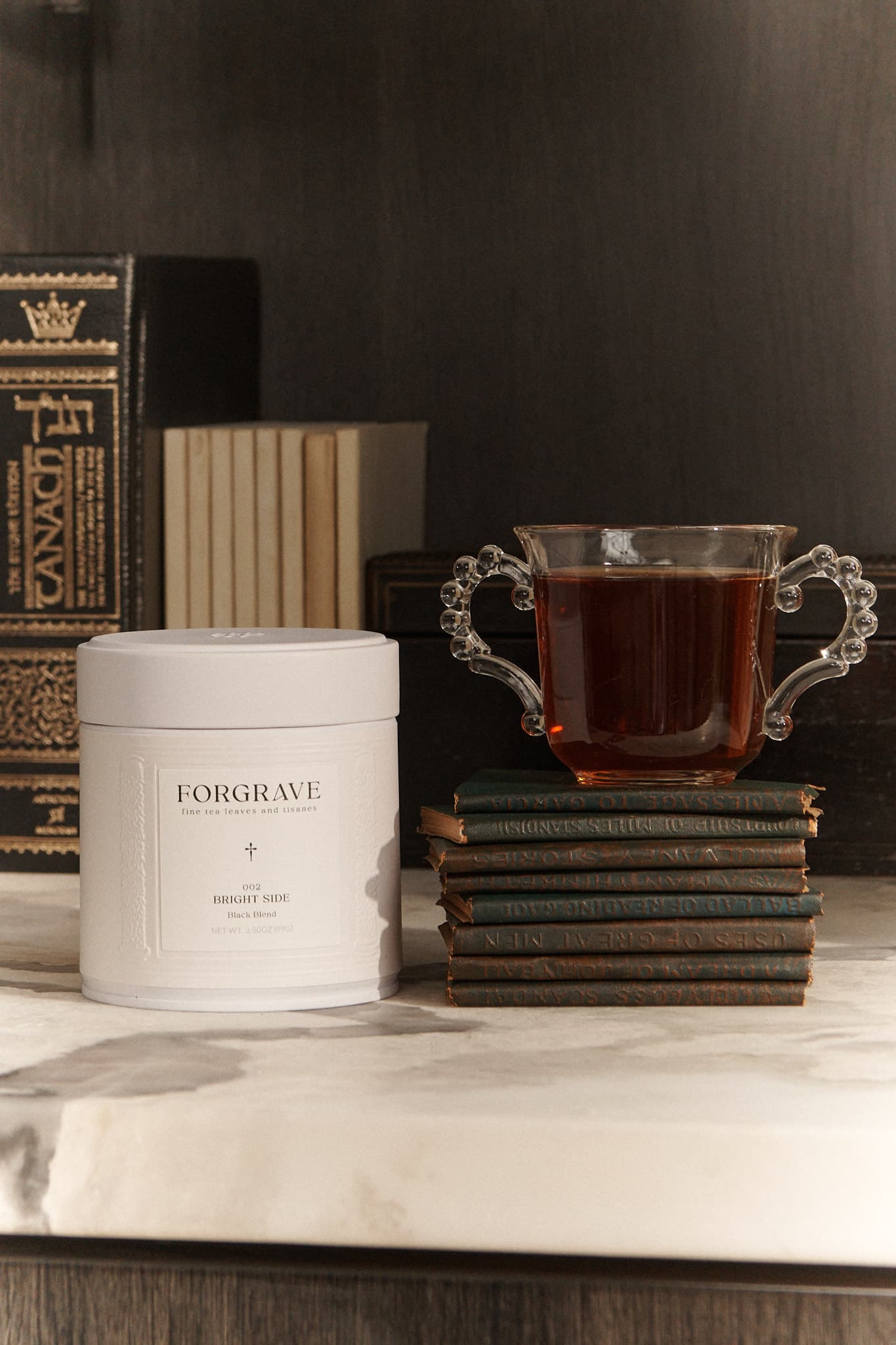FORGRAVE ‘Bright Side’ tea tin beside a glass cup of tea resting on stacked vintage books, styled on a marble surface.