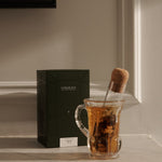 Glass mug with tea and a corked ‘Glass Cigar’ infuser beside a green FORGRAVE ‘Bright Side’ box on a marble shelf.