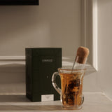 Glass mug with tea and a corked ‘Glass Cigar’ infuser beside a green FORGRAVE ‘Bright Side’ box on a marble shelf.