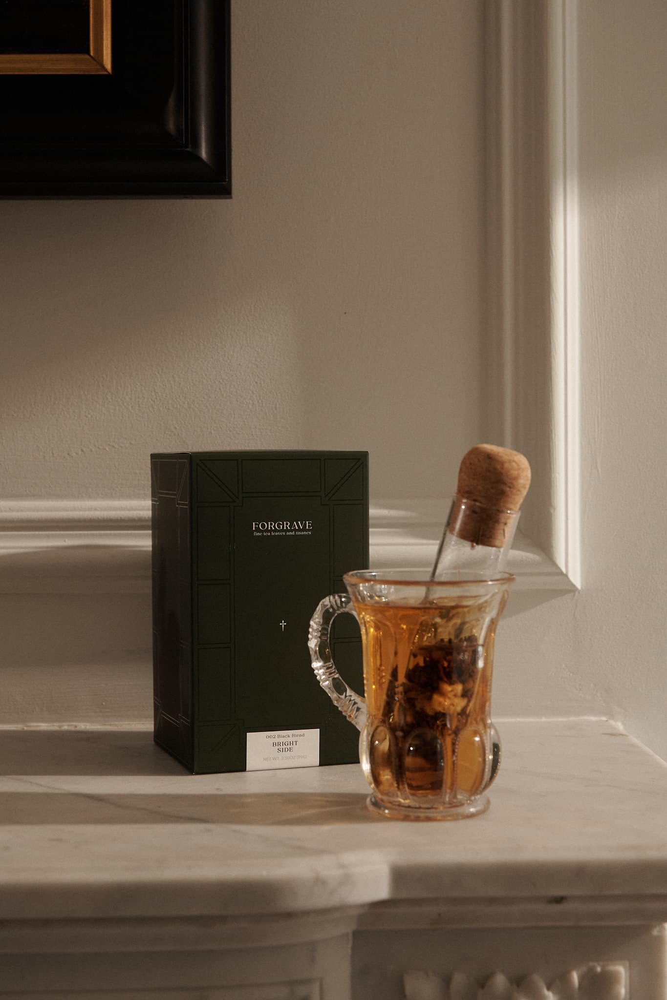 Glass mug with tea and a corked ‘Glass Cigar’ infuser beside a green FORGRAVE ‘Bright Side’ box on a marble shelf.