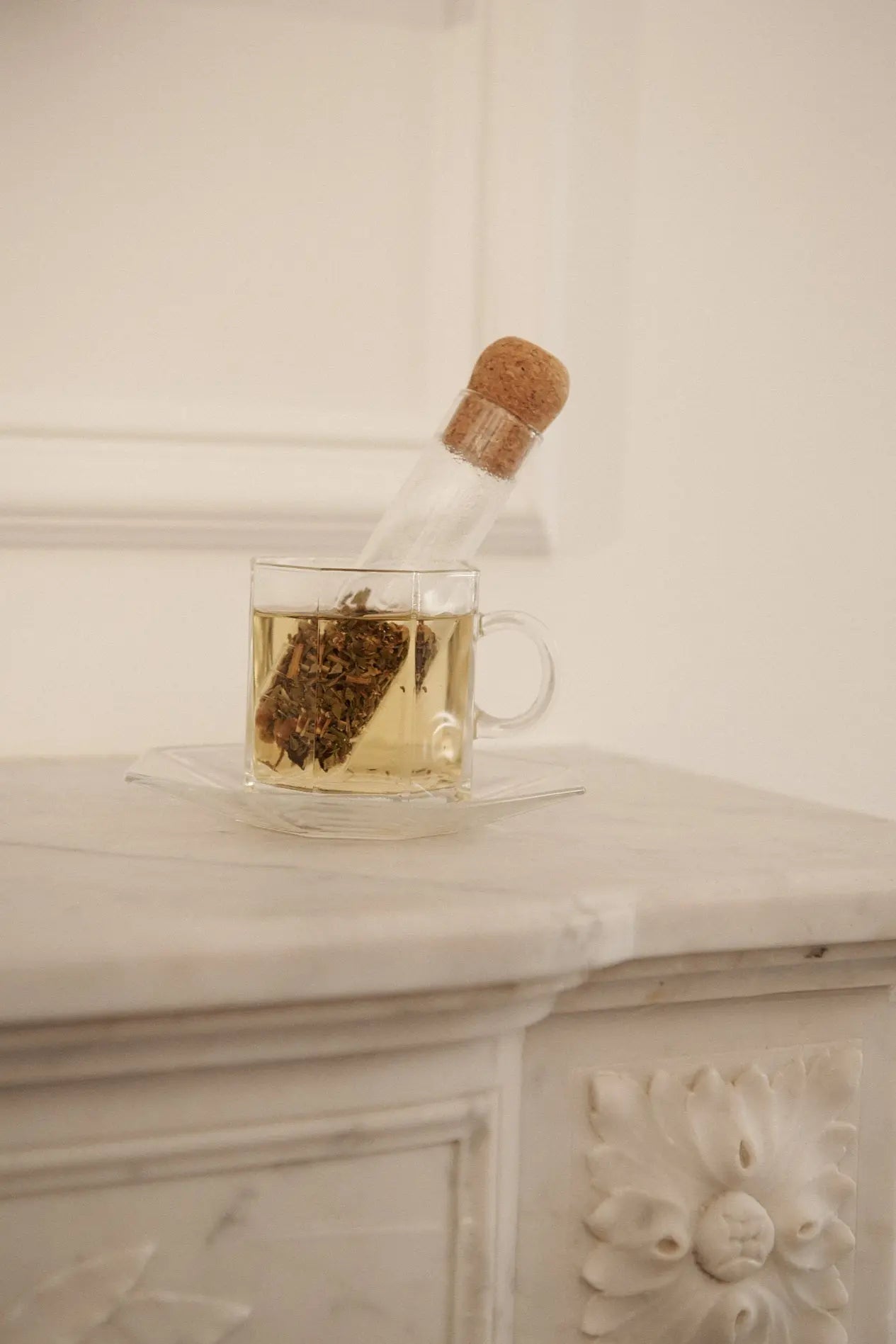 Clear glass teacup filled with steeped tea, with a cork-lidded glass infuser resting inside, placed on a white marble surface.