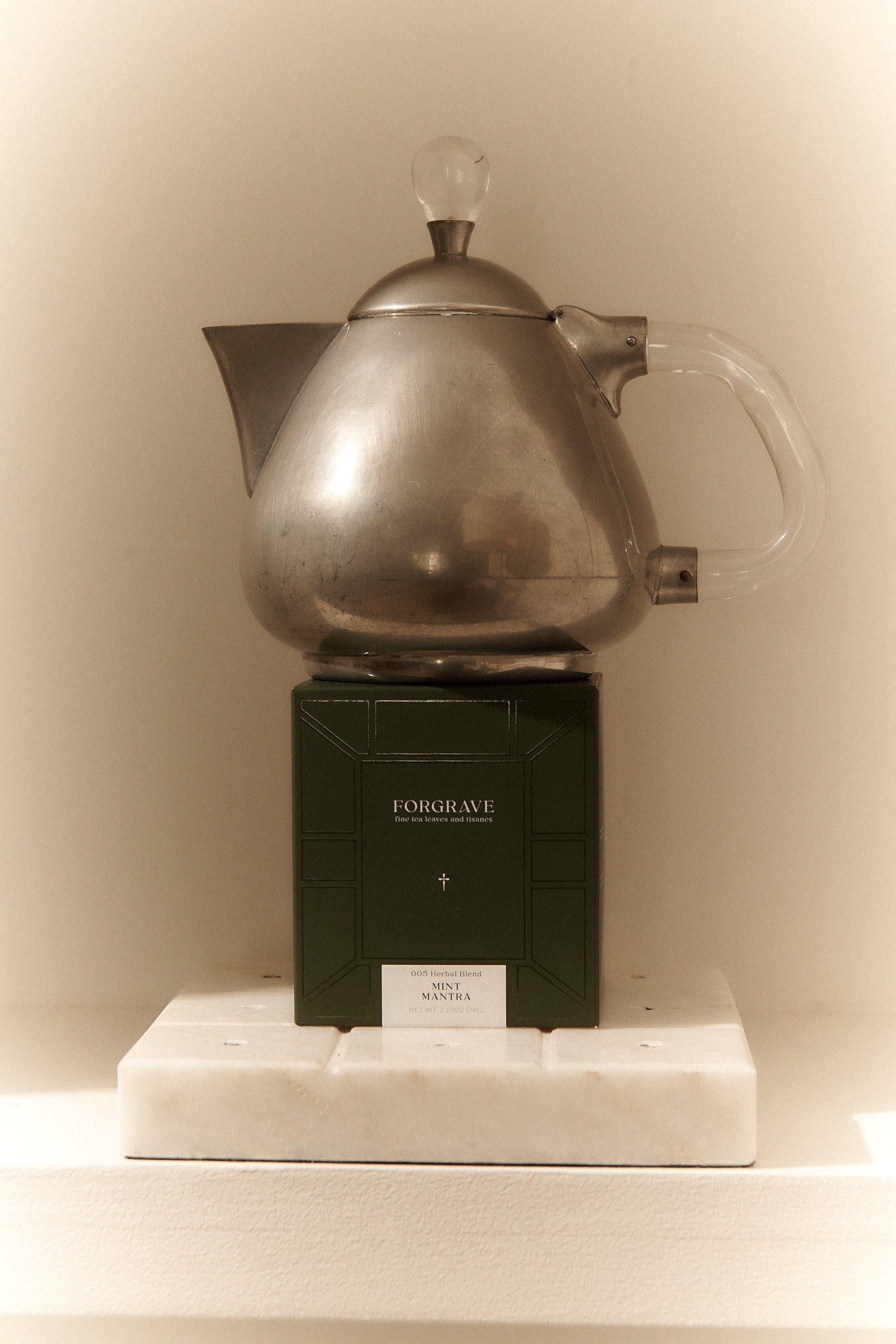 A metallic teapot with a clear handle sits atop a green tea box labeled “FORGRAVE Mint Mantra,” resting on a white marble base.