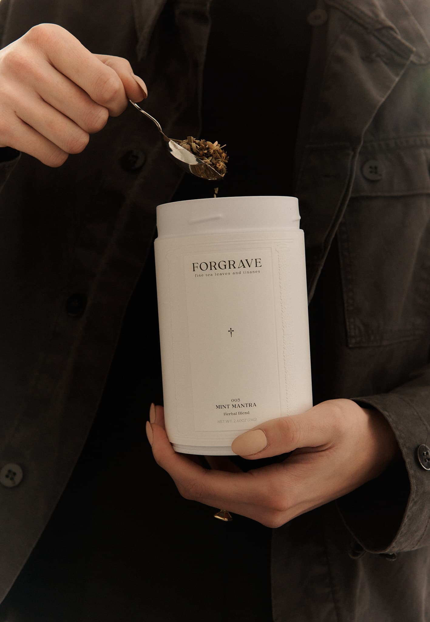 Person in black jacket scooping tea from a white ‘FORGRAVE 005 Mint Mantra white blend’ canister.