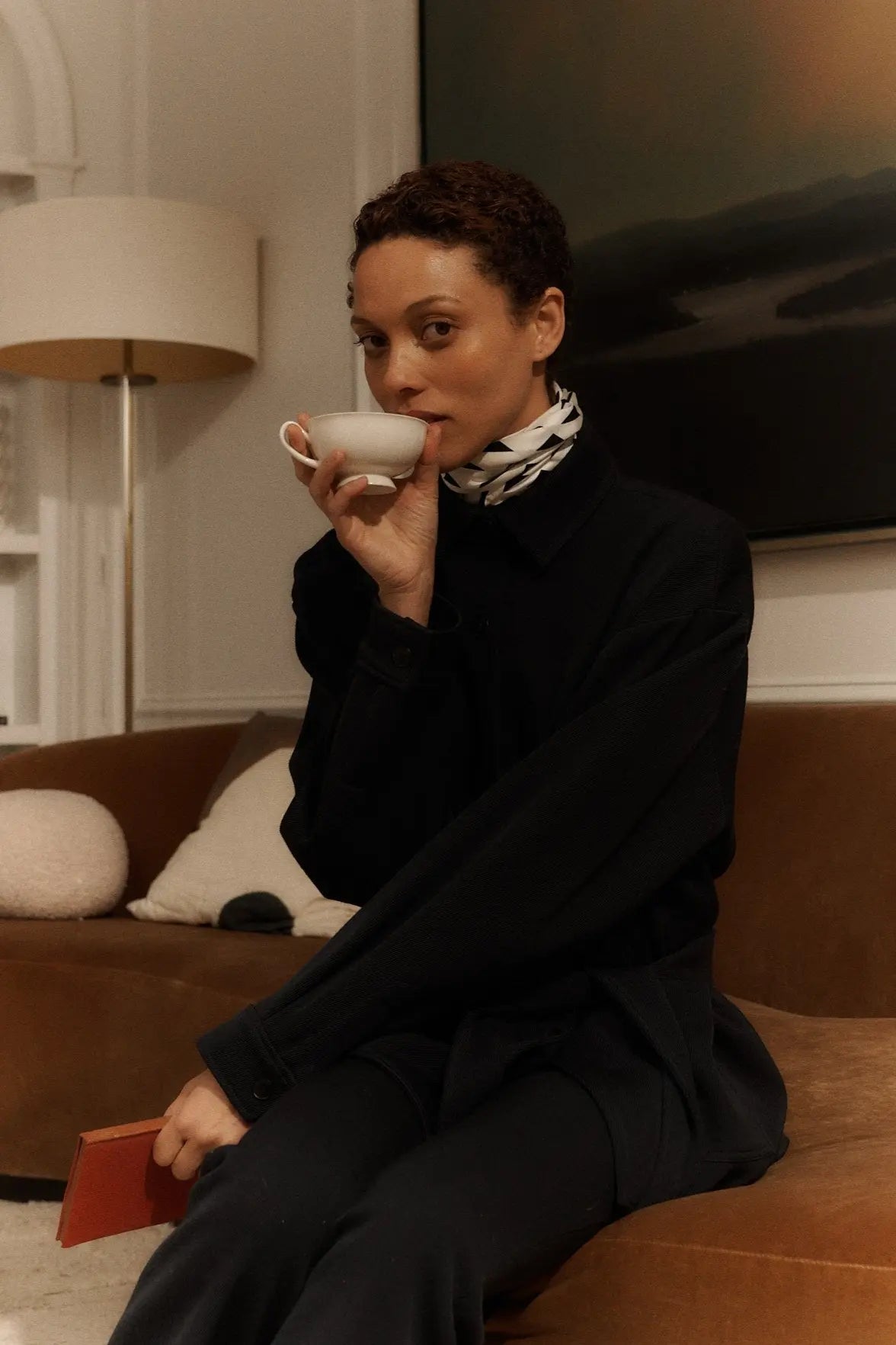 Person in a black outfit holding a white teacup, seated on a brown couch with a lamp and abstract artwork in the background.