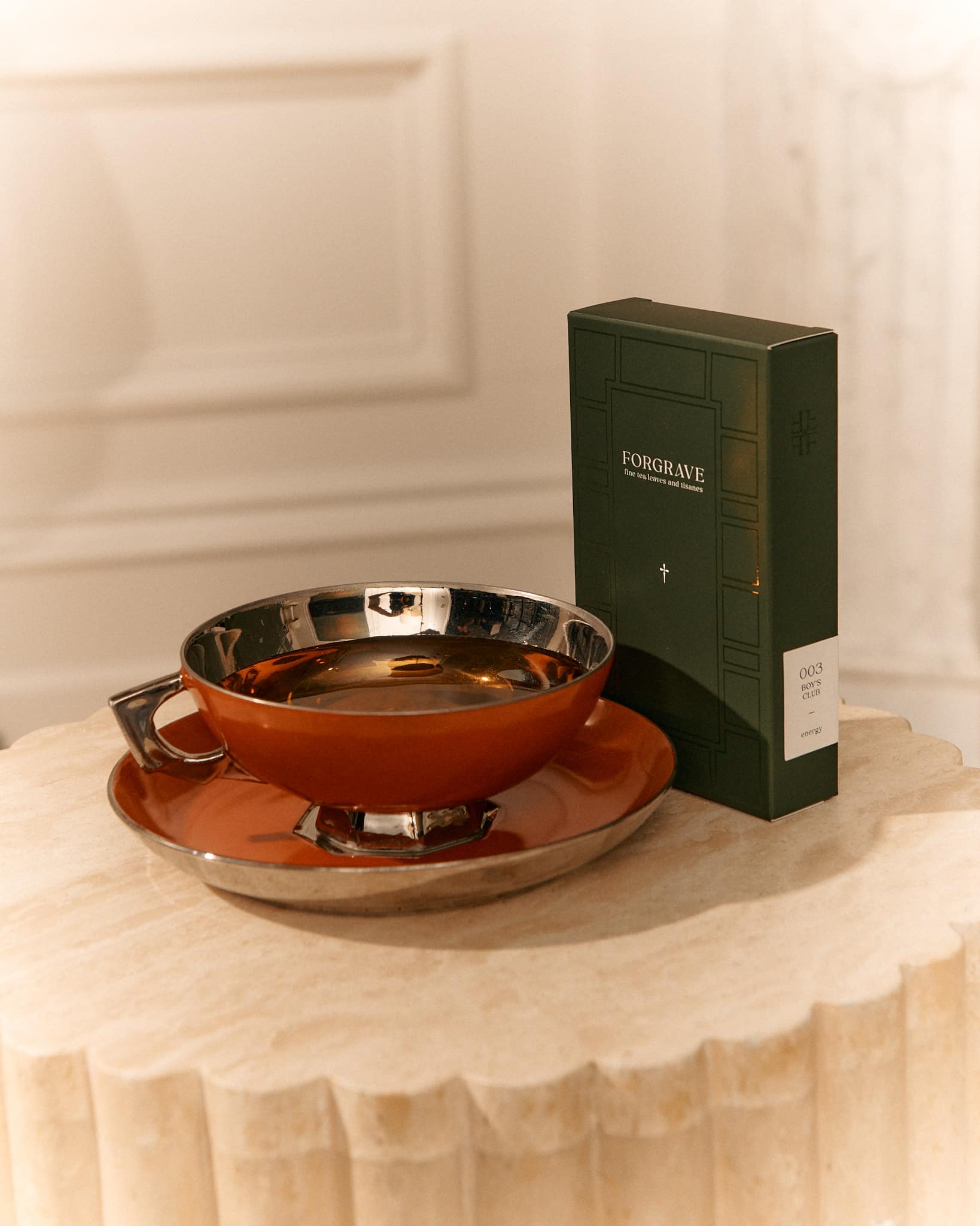A green Forgrave tea box labeled "003 Boys' Club" sits beside a metallic orange teacup and saucer on a textured beige table.