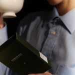 A person holds a white Forgrave "005 Mint Mantra Herbal Blend" box, reading the back label while holding a teacup, almost ready to drink.

