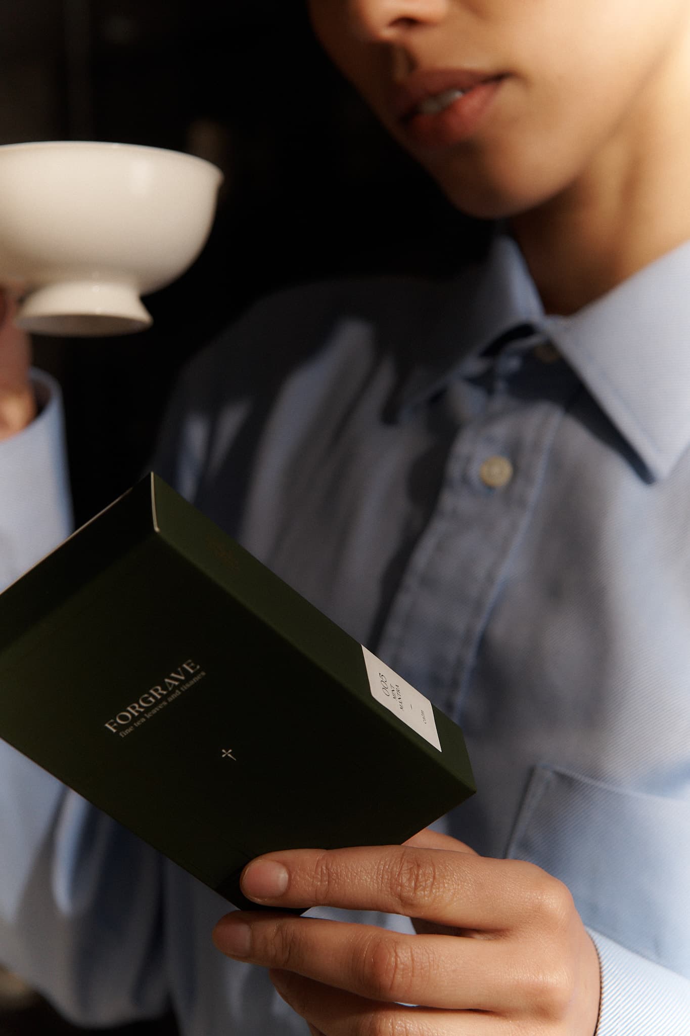A person holds a white Forgrave "005 Mint Mantra Herbal Blend" box, reading the back label while holding a teacup, almost ready to drink.

