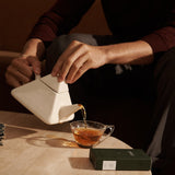 Person pours tea from a modern white teapot into a glass mug, with a FORGRAVE ‘Bright Side’ box on a stone table nearby.