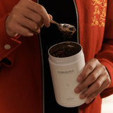 Person scooping tea from a white Forgrave "003 Boys' Club" canister, wearing a red jacket with gold embroidery on the chest.