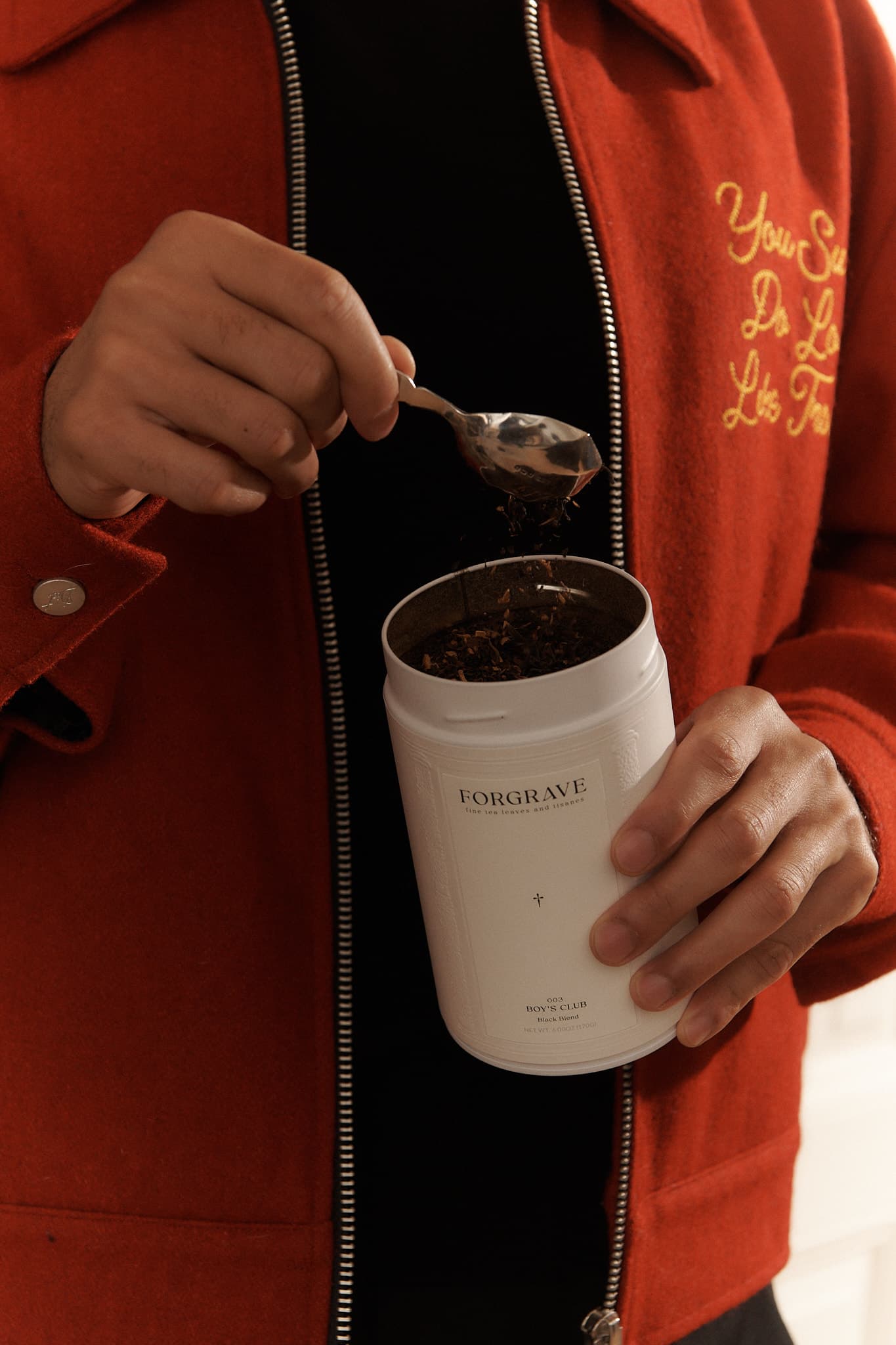 Person scooping tea from a white Forgrave "003 Boys' Club" canister, wearing a red jacket with gold embroidery on the chest.