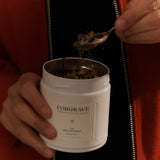 A person in a red jacket holding a white tea canister labeled ‘FORGRAVE 005 Mint Mantra Herbal Blend,’ scooping tea with a spoon.