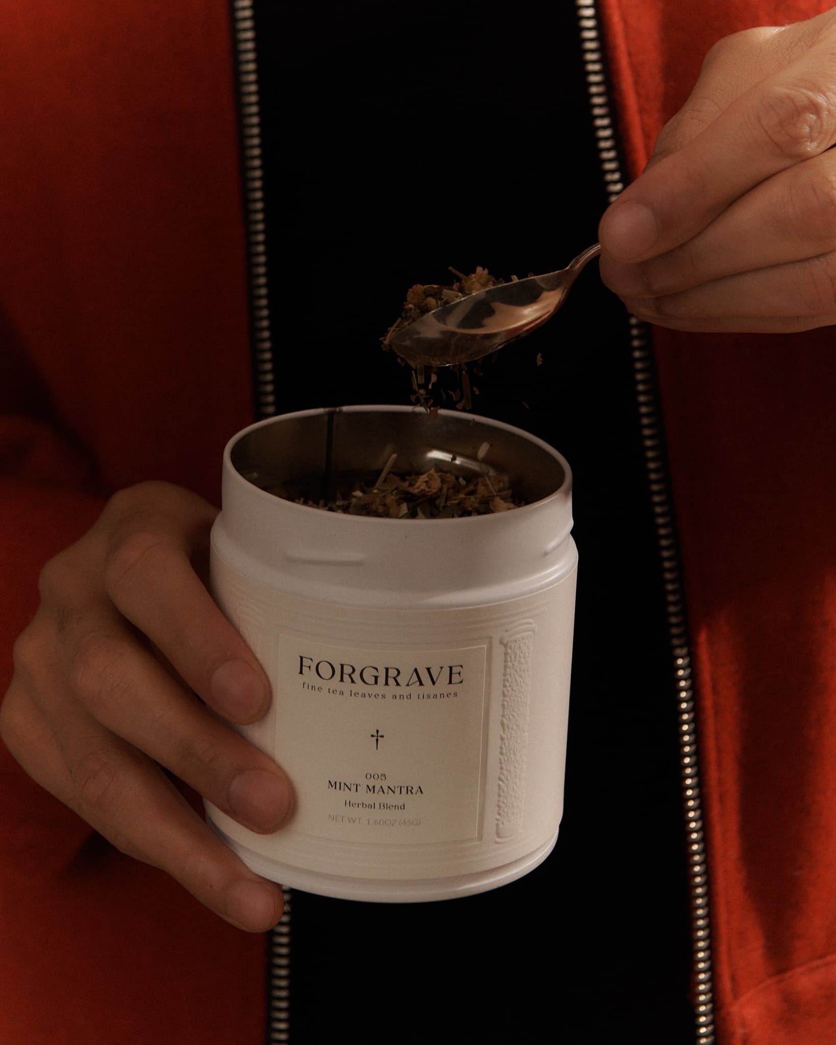 A person in a red jacket holding a white tea canister labeled ‘FORGRAVE 005 Mint Mantra Herbal Blend,’ scooping tea with a spoon.