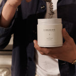 Person scooping tea from a white Forgrave "003 Boys' Club" canister, wearing a navy jacket and a white shirt.