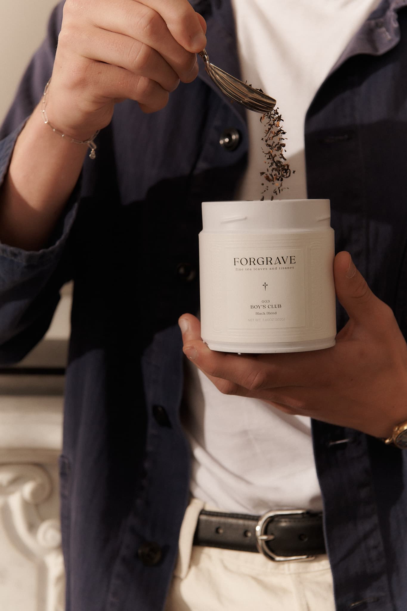 Person scooping tea from a white Forgrave "003 Boys' Club" canister, wearing a navy jacket and a white shirt.
