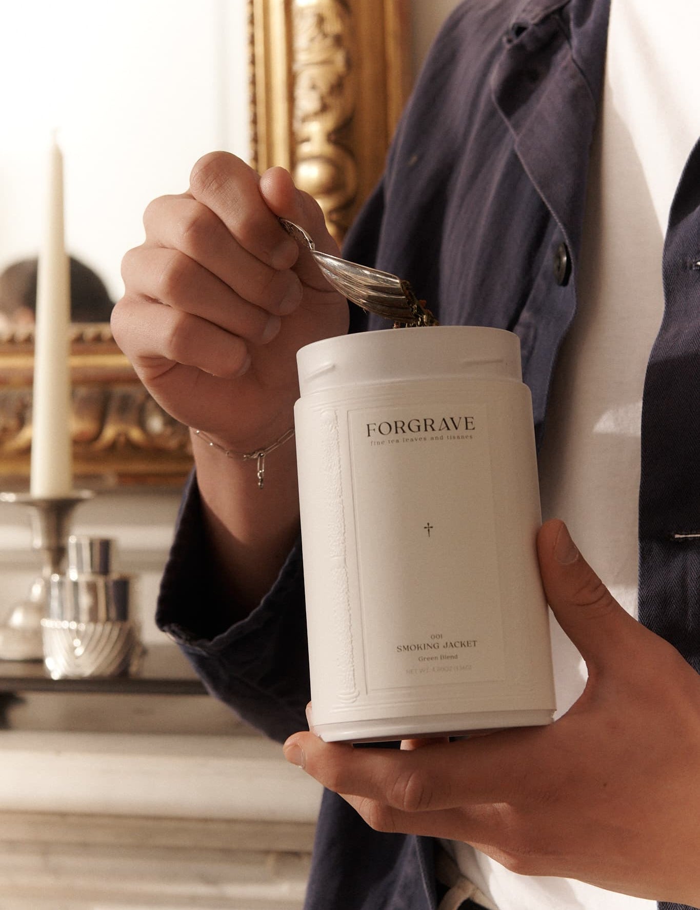 Person scooping tea from a white ‘FORGRAVE Smoking Jacket Green Blend’ canister, with a gold-framed mirror and candle in the background.