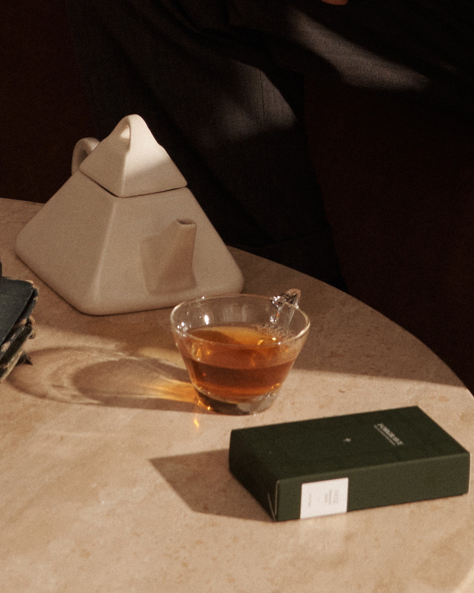 A modern ceramic teapot, a glass teacup with amber tea, and a green tea box sit on a beige table in warm, natural lighting.