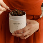 A person in an orange outfit holding a white tea canister labeled ‘FORGRAVE 001 Smoking Jacket Green Blend,’ scooping tea with a spoon.