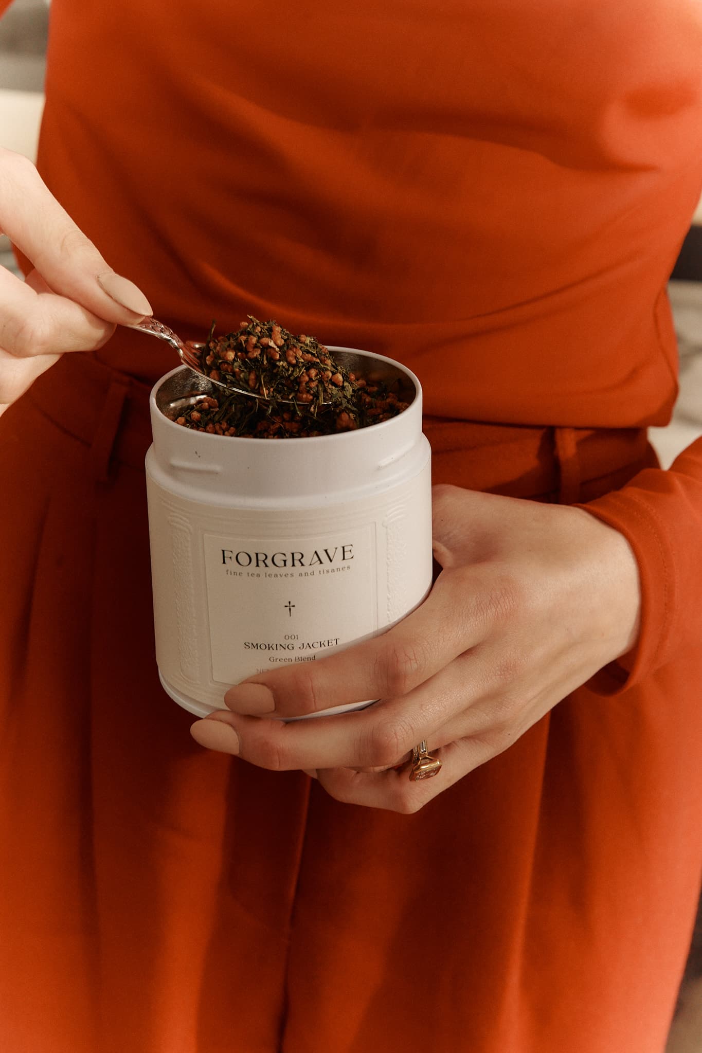 A person in an orange outfit holding a white tea canister labeled ‘FORGRAVE 001 Smoking Jacket Green Blend,’ scooping tea with a spoon.