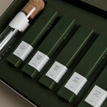 Open green box containing five dark green tea tubes labeled ‘001 Smoking Jacket’ to ‘005 Mint Mantra,’ with a glass tea infuser.