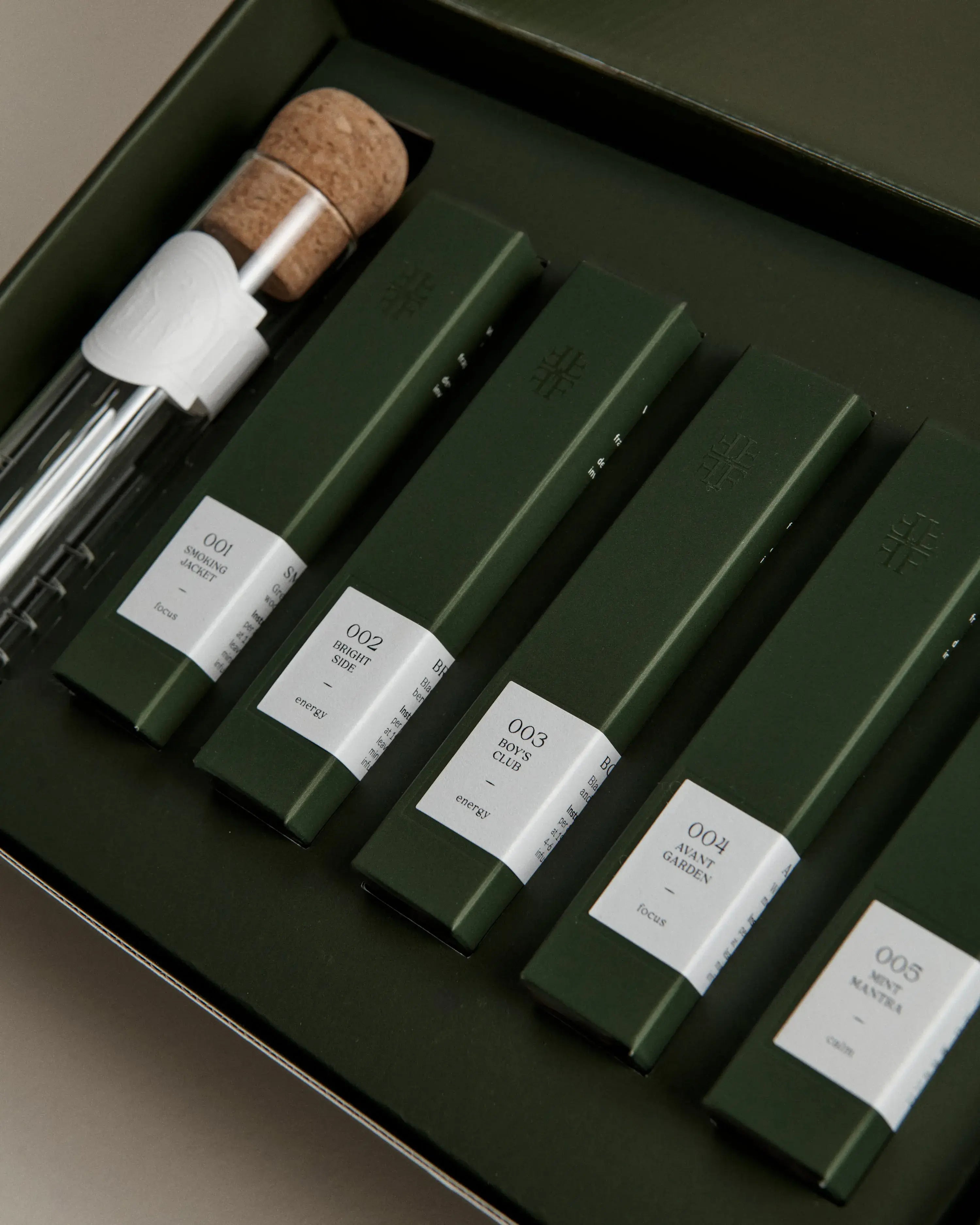 Open green box containing five dark green tea tubes labeled ‘001 Smoking Jacket’ to ‘005 Mint Mantra,’ with a glass tea infuser.