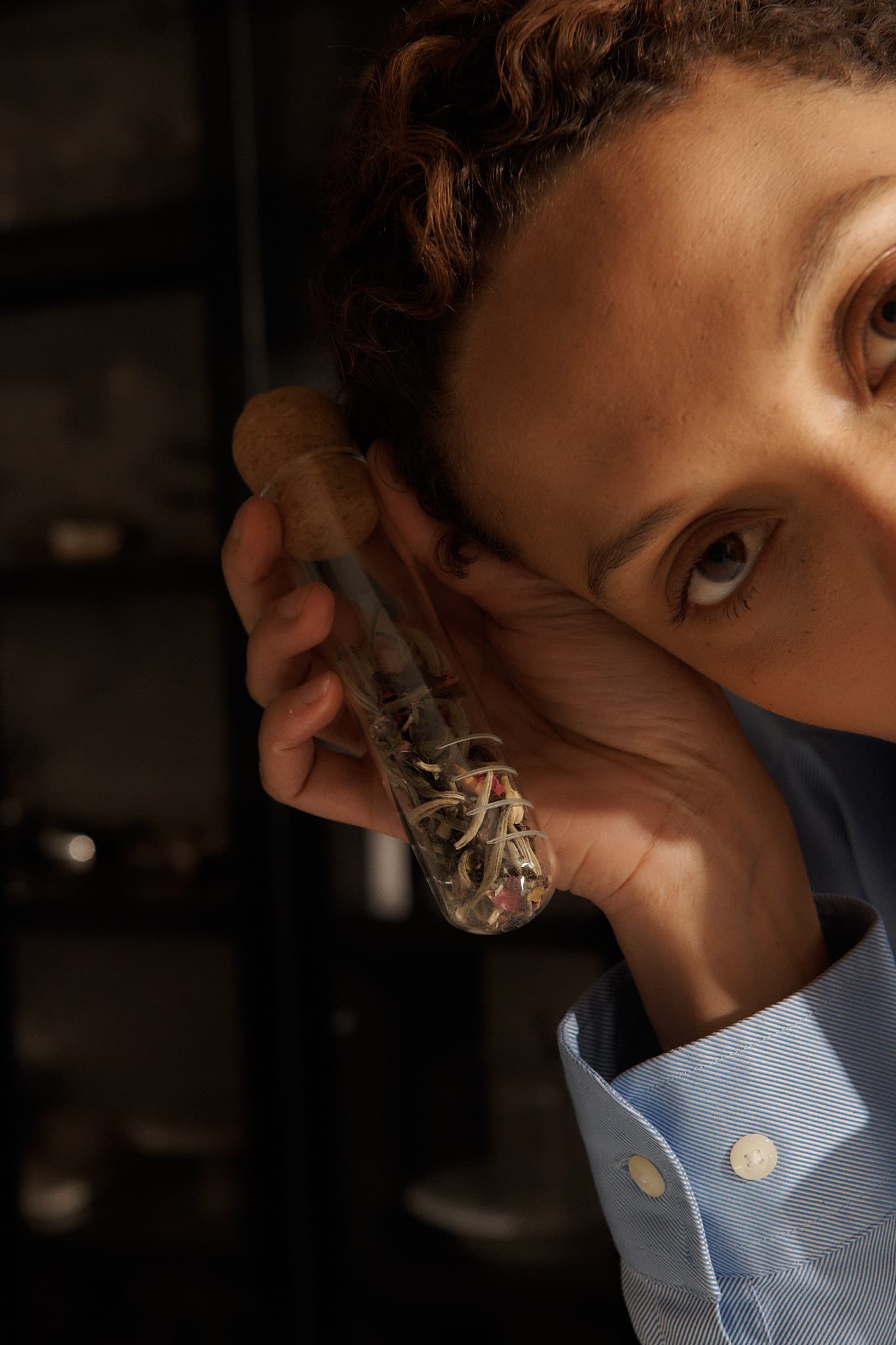 Hand holding a glass tea infuser tube with a cork lid, filled with loose tea leaves, against a dimly lit background.
