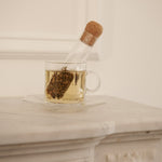 Clear glass teacup filled with steeped tea, with a cork-lidded glass infuser resting inside, placed on a white marble surface.