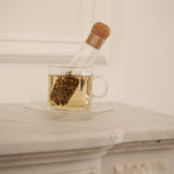 Clear glass teacup filled with steeped tea, with a cork-lidded glass infuser resting inside, placed on a white marble surface.