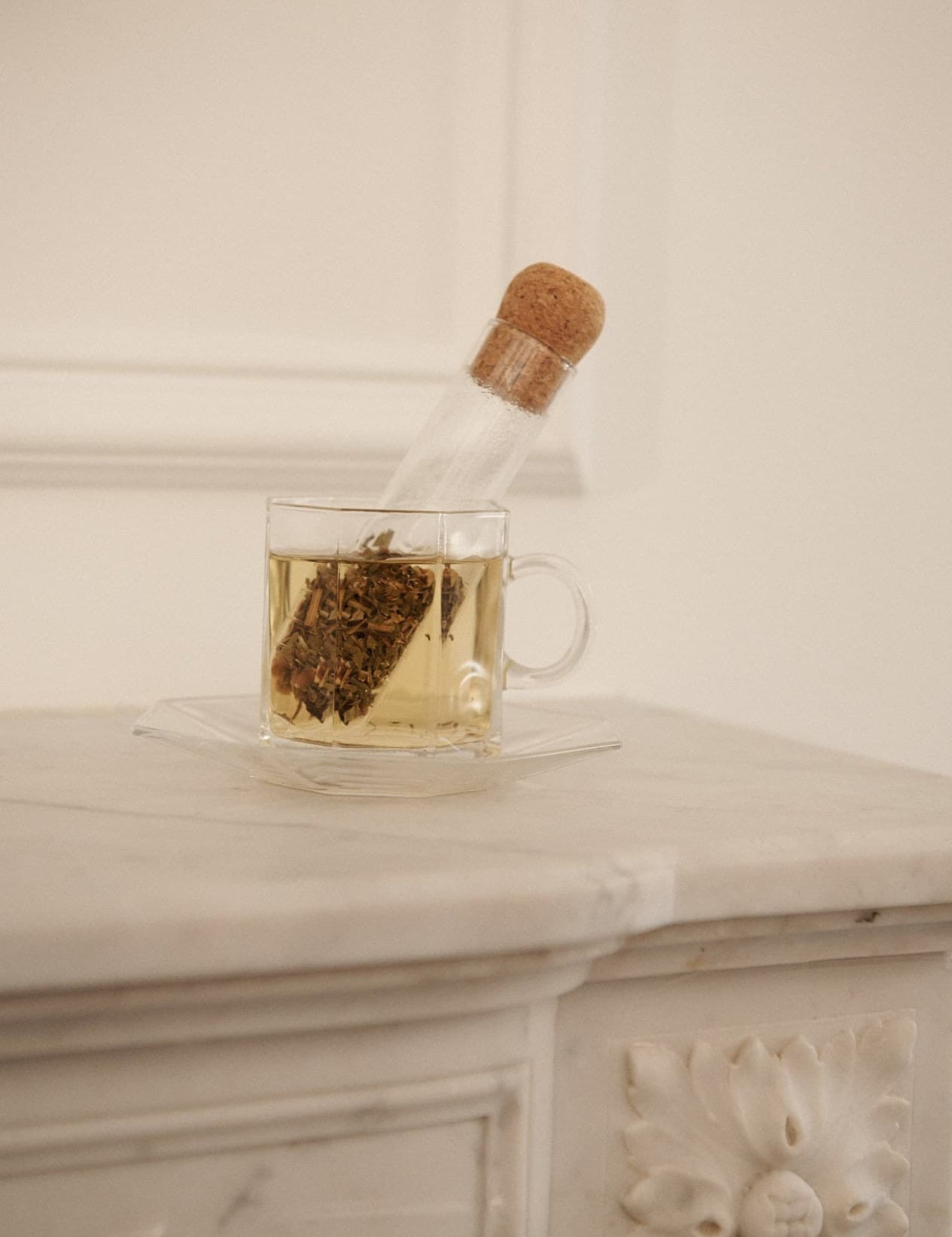 Clear glass teacup filled with steeped tea, with a cork-lidded glass infuser resting inside, placed on a white marble surface.