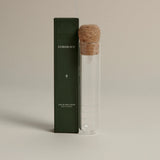 Dark green box labeled ‘FORGRAVE The Glass Cigar Fine Tea Infuser’ next to a clear glass infuser tube with a cork lid on a beige background.