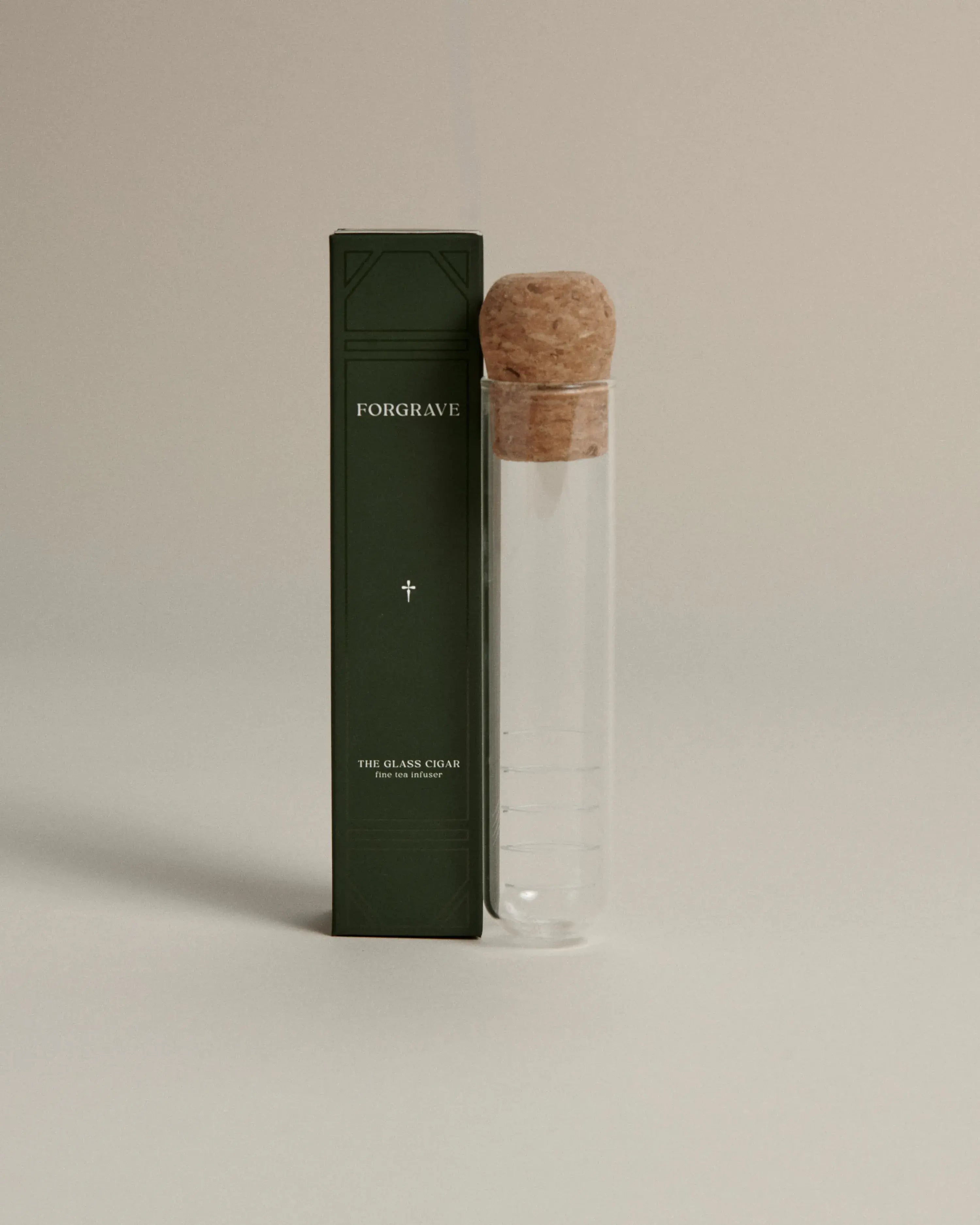 Dark green box labeled ‘FORGRAVE The Glass Cigar Fine Tea Infuser’ next to a clear glass infuser tube with a cork lid on a beige background.