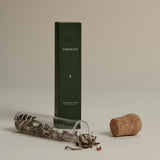 Dark green box labeled ‘FORGRAVE The Glass Cigar’ next to a cork and a glass tube spilling loose tea leaves with floral accents.