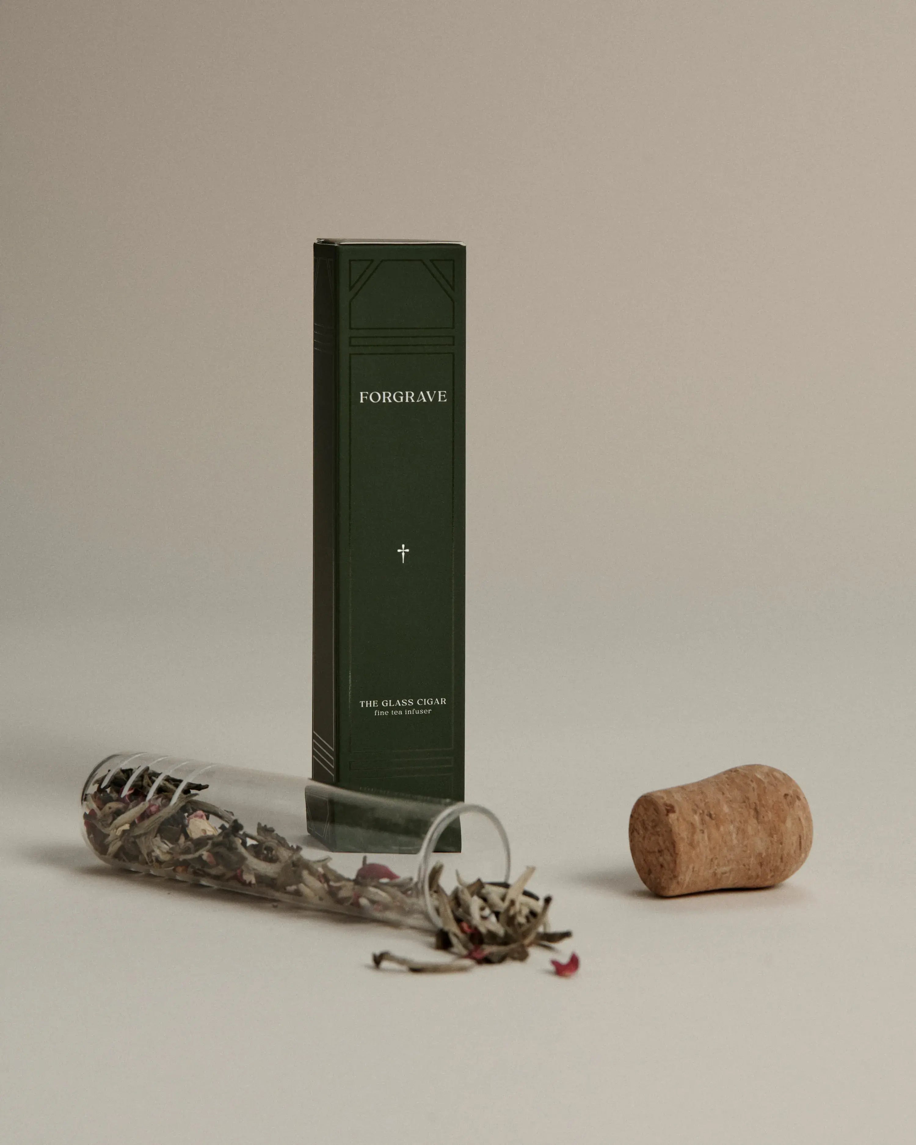 Dark green box labeled ‘FORGRAVE The Glass Cigar’ next to a cork and a glass tube spilling loose tea leaves with floral accents.
